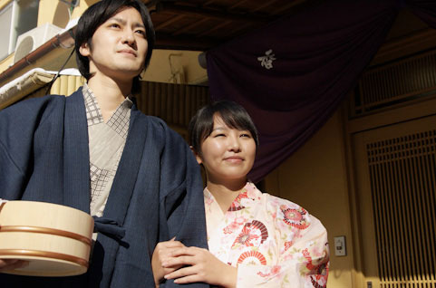 温泉に向かうカップルを撮影したフリー写真。浴衣を着て風呂桶を持った姿が日本的。